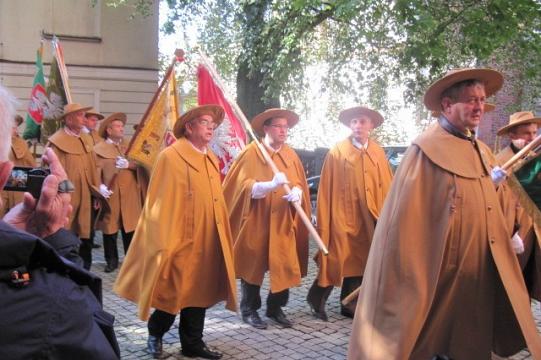 Barwny pochód pszczelarzy w złotych pelerynach, niosących flagi podczas uroczystości.