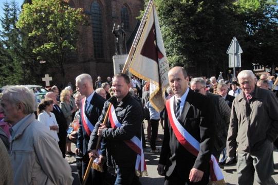 Uroczysty pochód pszczelarzy w garniturach z szarfami i flagą podczas obchodów.