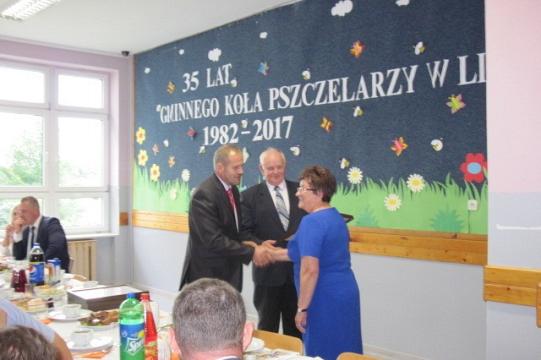 Dwie osoby ściskają dłonie na tle dekoracji 35-lecia Koła Pszczelarzy w Linii.