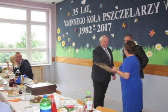 Uczestnicy 35-lecia Koła Pszczelarzy wymieniają uściski dłoni na tle okolicznościowej dekoracji.