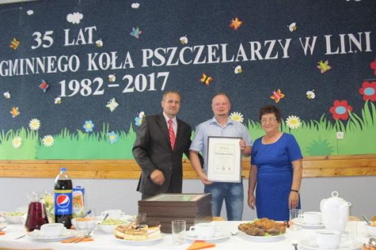 Trzy osoby pozują z nagrodą przed banerem upamiętniającym 35-lecie Koła Pszczelarzy.