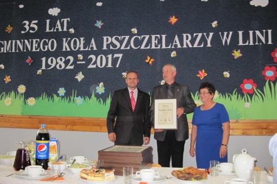 Trzy osoby pozują przed banerem 35-lecia Koła Pszczelarzy w Lini, odbierając wyróżnienie.