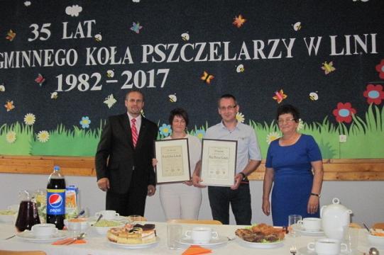 Cztery osoby na tle banera 35-lecia Koła Pszczelarzy w Lini, trzymające dokumenty.
