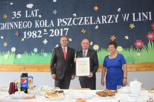 Trzy osoby z nagrodą przed transparentem 35-lecia Koła Pszczelarzy w Linii.
