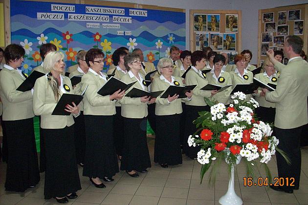 Chór Pięciolinia śpiewa z nutami, dyrygowany przez osobę z prawej.