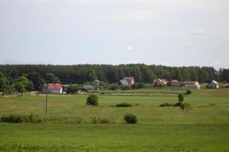 Malowniczy widok Pobłocia z zielonymi polami, zabudowaniami i lasem w tle.