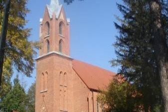 Ceglany kościół w Smażynie z wieżą i spiczastym dachem, otoczony drzewami na tle nieba.