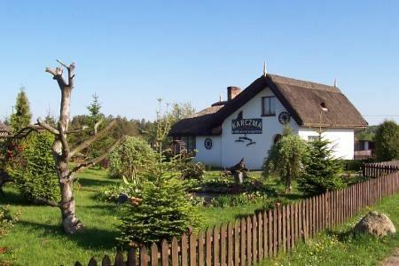 Biały dom z brązowym dachem, otoczony zielonym ogrodem i drewnianym płotem, pod błękitnym niebem.