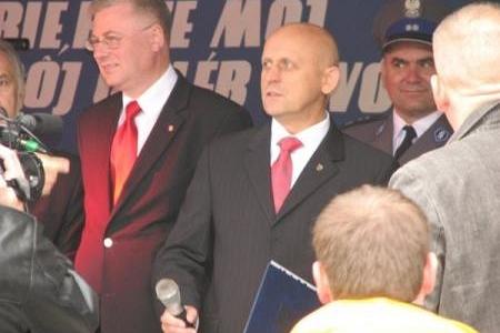 Dwóch mężczyzn w garniturach, z banerem w tle, podczas Dożynek Powiatowo-Gminnych.