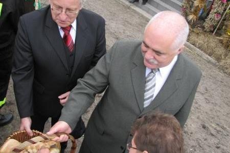 Dwóch mężczyzn w garniturach na dożynkach, jeden odbiera chleb.