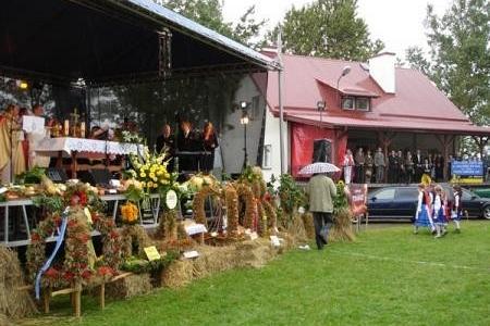 Scena z dożynek gminnych 2008 z drewnianym stoiskiem i czerwonym budynkiem na tle zielonego trawnika.