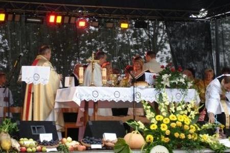 Scena dożynkowa z księdzem, ołtarzem, ozdobami z plonów i kwiatami podczas uroczystości.