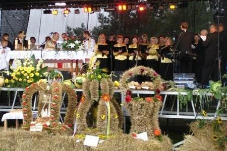Duże, ozdobne słomiane wieńce dożynkowe i ludzie na uroczystości gminnej.