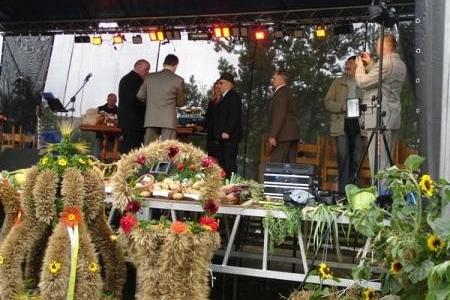 Wieńce dożynkowe na pierwszym planie, w tle scena z ludźmi podczas Dożynek Gminnych.