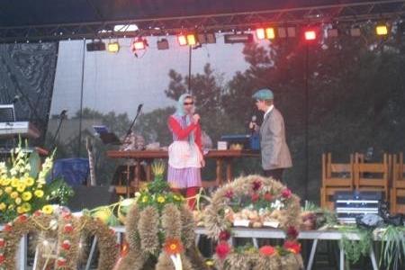 Dożynki: Kobieta i mężczyzna w strojach ludowych na scenie, wokół stoły z plonami.