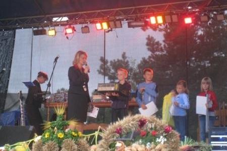 Scena dożynkowa z występem muzycznym, prowadzącą, chórem dziecięcym i wieńcami.