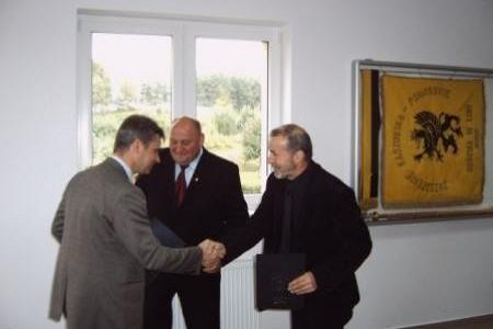 Oficjalny uścisk dłoni trzech mężczyzn w garniturach, w tle okno i dekoracja ścienna.