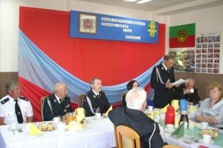 Uczestnicy Zjazdu OSP przy stołach, jedna osoba przemawia na tle dekoracji.