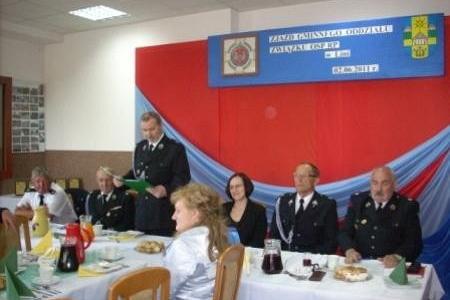Zjazd OSP z uczestnikami w mundurach przy stołach, ze stojącym mówcą i banerem w tle.