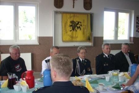 Uczestnicy Zjazdu OSP 2011 siedzą przy stole, w tle żółty sztandar i okna.