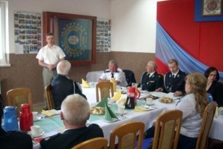 Uczestnicy Zjazdu OSP 2011 siedzą przy stole, słuchając prelegenta na tle flagi.