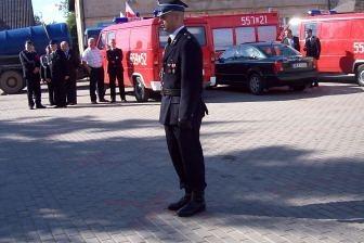 Strażak OSP w mundurze na placu, w tle czerwone wozy strażackie i budynki.