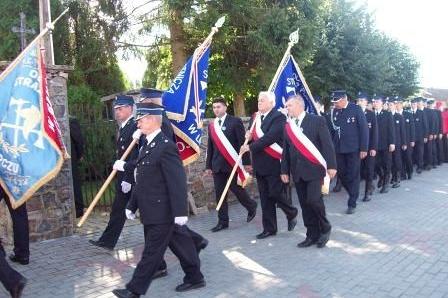 Strażacy z OSP Lewino maszerują w uroczystym pochodzie ze sztandarami i flagami.
