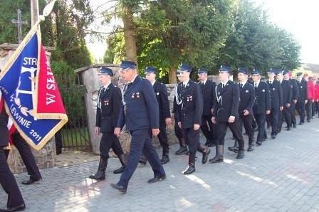 Poczet sztandarowy i strażacy OSP Lewino maszerują w uroczystym pochodzie.