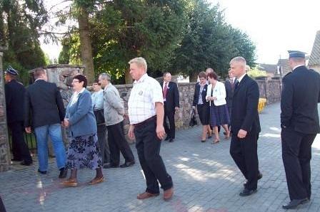 Grupa ludzi w formalnych strojach, w tym ksiądz, uczestniczy w uroczystości OSP Lewino.