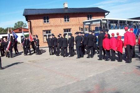 Członkowie OSP Lewino w mundurach i czerwonych kurtkach przed remizą z wozem strażackim.