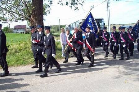 Strażacy OSP w galowych mundurach niosą sztandary podczas uroczystego marszu.