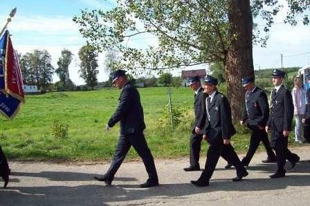 Pochód mężczyzn w garniturach ze sztandarem OSP Lewino na tle zieleni.
