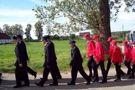 Dwie grupy osób maszerują: strażacy OSP Lewino w czerwonych strojach i osoby w ciemnych.