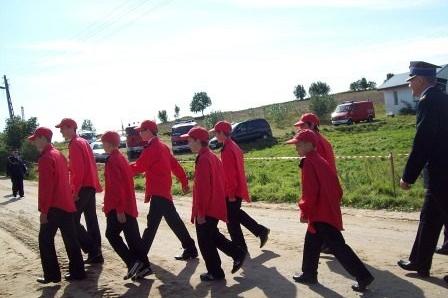 Młodzi strażacy OSP Lewino w czerwonych kurtkach maszerują w słoneczny dzień.