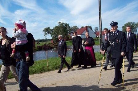 Pochód strażaków OSP Lewino z duchownymi nad wodą w słoneczny dzień.