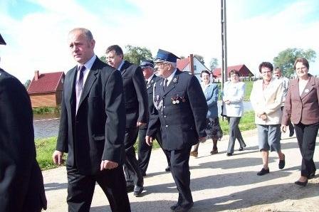 Mężczyźni w garniturach i mundurach OSP idą ulicą w słoneczny dzień.