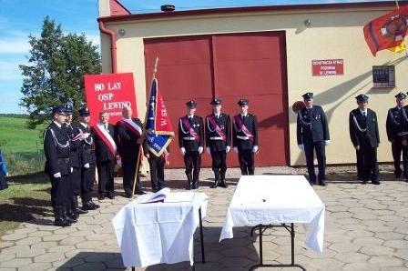 Strażacy OSP Lewino w formacji przed remizą, uczestniczący w ceremonii ze sztandarem.