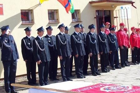 Uroczystość OSP Lewino: strażacy w szyku i osoby w czerwonych strojach przed budynkiem.