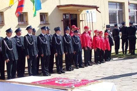Strażacy OSP Lewino i młodzież w mundurach podczas apelu przed budynkiem udekorowanym flagami.