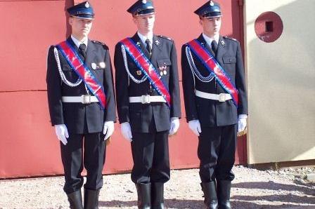 Trzech członków OSP Lewino w galowych mundurach z szarfami stoi na baczność.