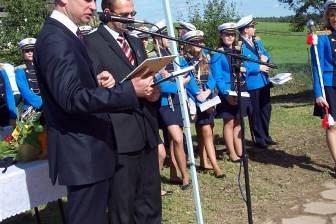 Mężczyźni w garniturach i strażacy OSP przy mikrofonie na uroczystości pod namiotem.