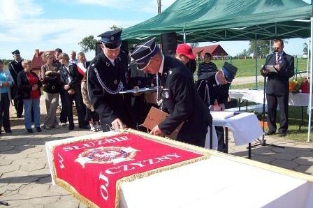 Nowy sztandar OSP Lewino na stole, obok strażacy podczas uroczystości.