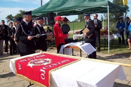 Czerwono-biały sztandar na stole podczas uroczystości OSP Lewino pod zielonym namiotem.