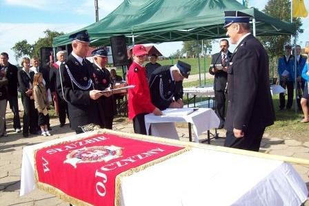 Uczestnicy uroczystości pod zielonym namiotem, obok stół z czerwonym sztandarem OSP Lewino.
