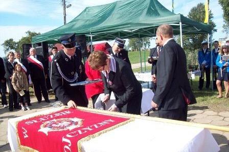 Ludzie w mundurach i garniturach pochylają się nad sztandarem OSP Lewino na ceremonii.
