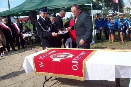 Sztandar OSP Lewino na stole, w tle mężczyźni i namiot podczas ceremonii.