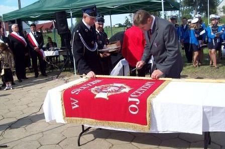 Mężczyźni przy stole oglądają czerwony sztandar OSP Lewino podczas uroczystości.