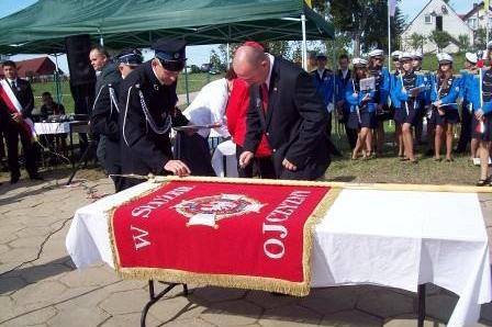 Uroczystość wręczenia sztandaru OSP Lewino z osobami przy stole i strażakami w tle.