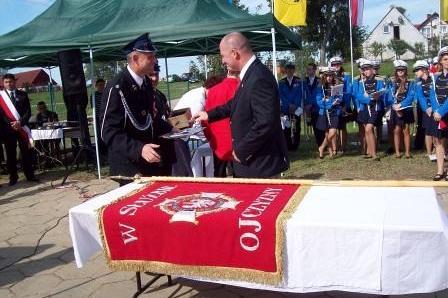 Wręczenie sztandaru OSP Lewino, mężczyźni ściskają dłonie, sztandar leży na stole.