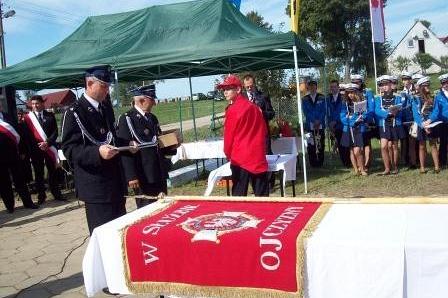 Uroczystość z czerwonym sztandarem OSP Lewino, w tle uczestnicy i zielony namiot.
